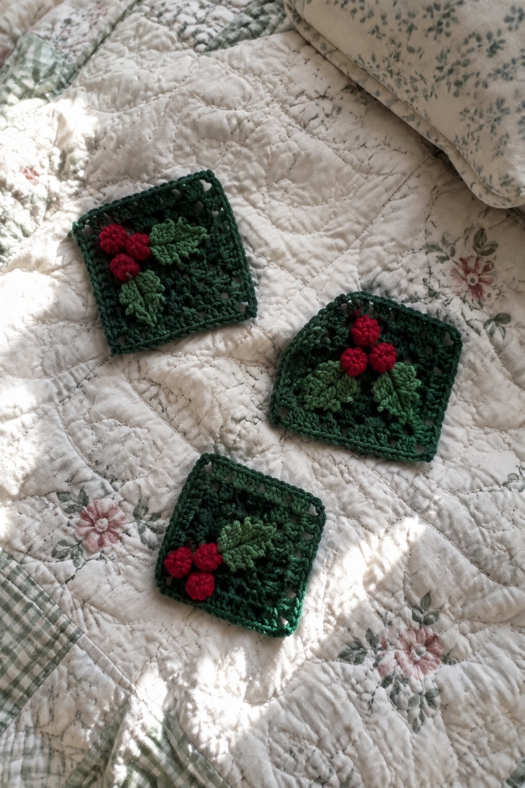 Three green crocheted holly squares with red berries on a floral quilt.