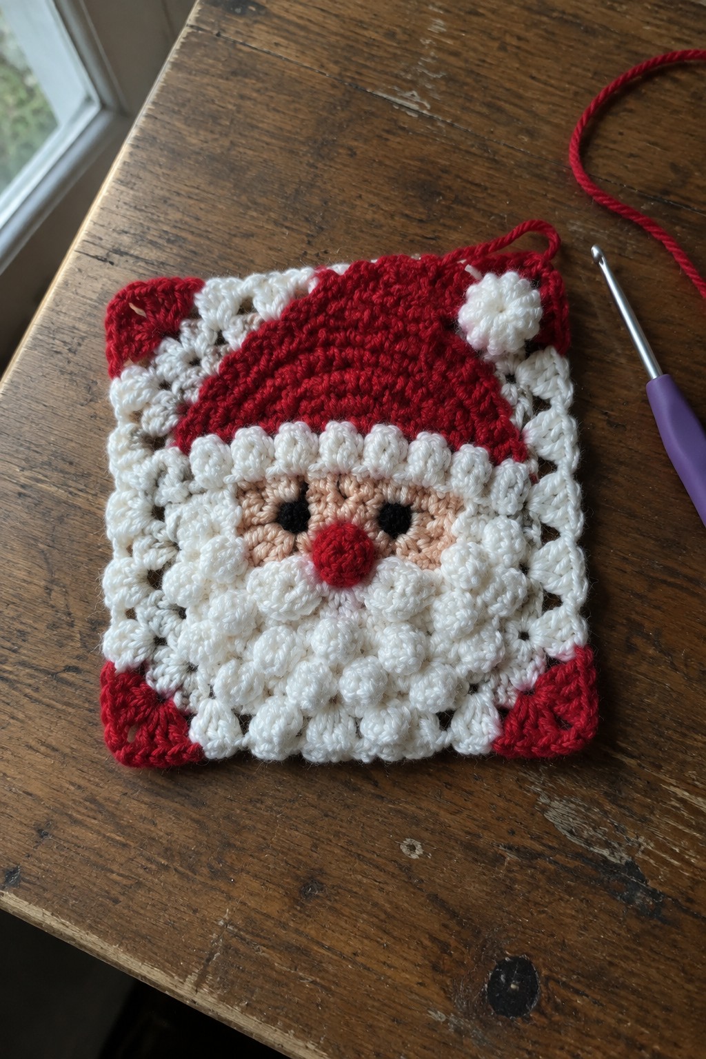 Crocheted Santa Claus square with red hat and beard beside crochet hook