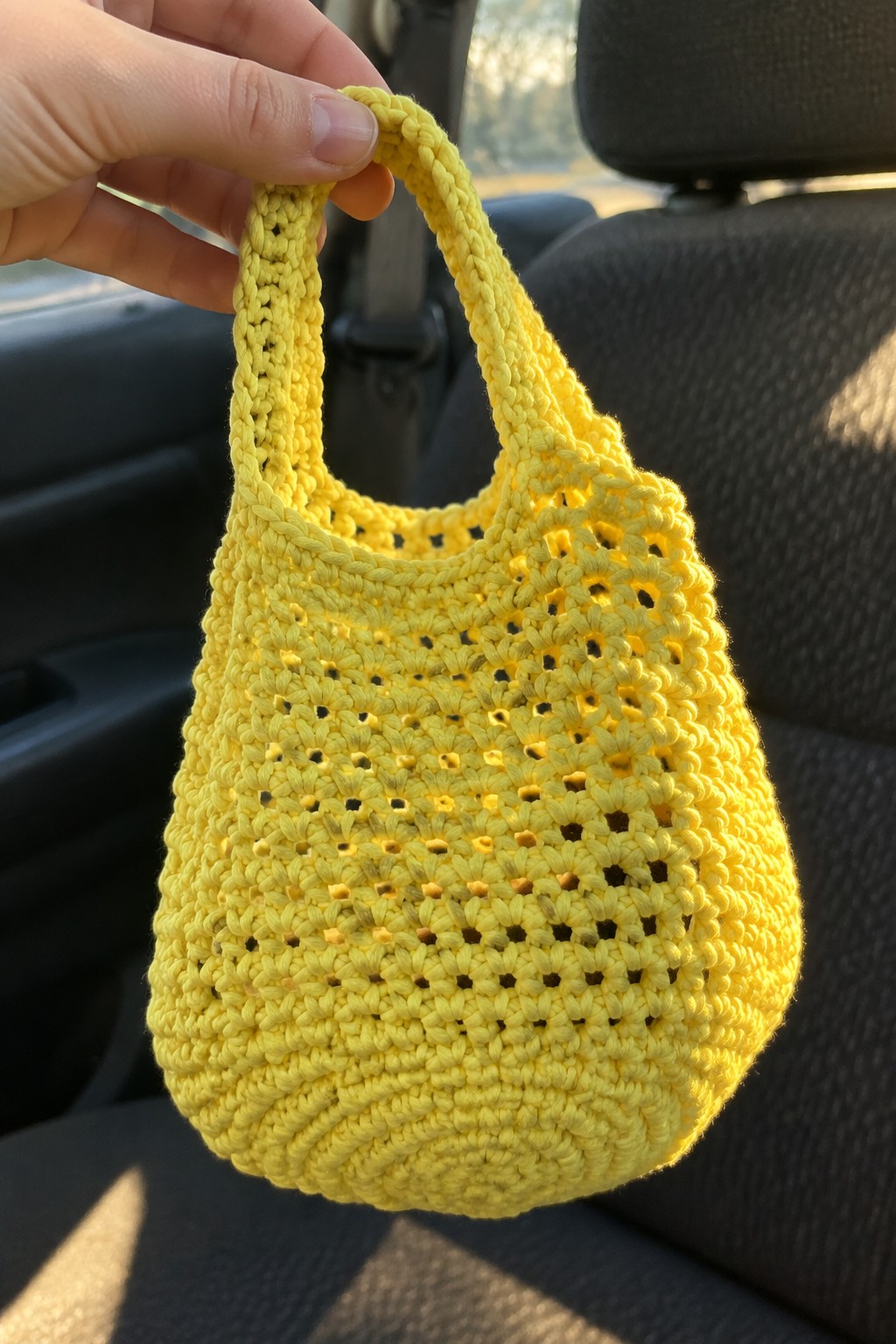 Hand holding yellow crocheted mesh bag inside sunlit car interior