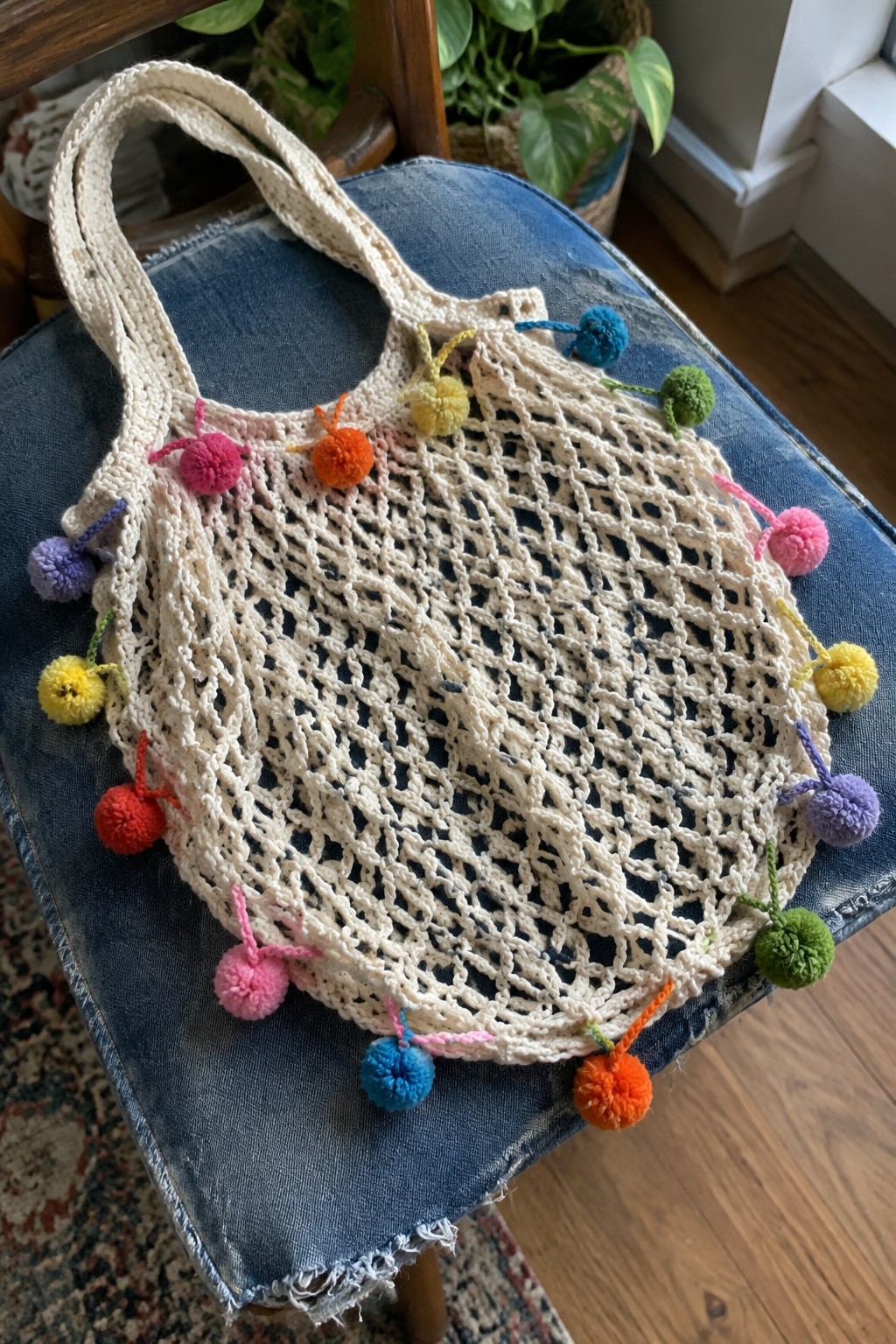 Cream crochet net bag with colorful pom-poms on blue denim chair.