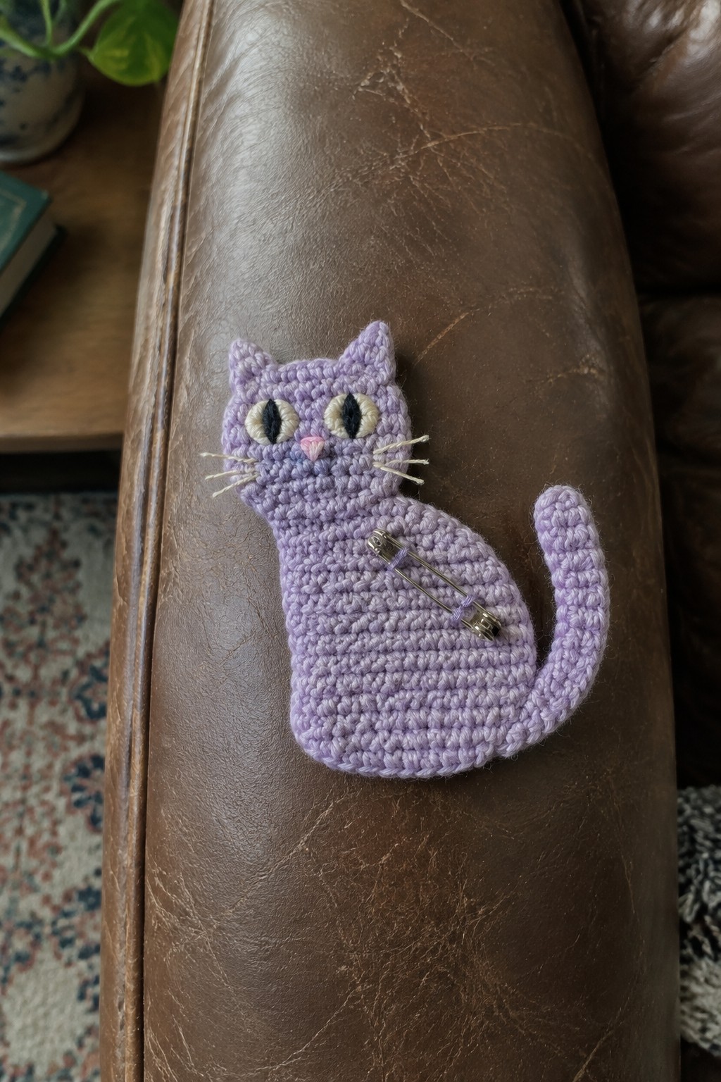 A purple crocheted cat brooch with embroidered face and safety pin back rests on brown leather.