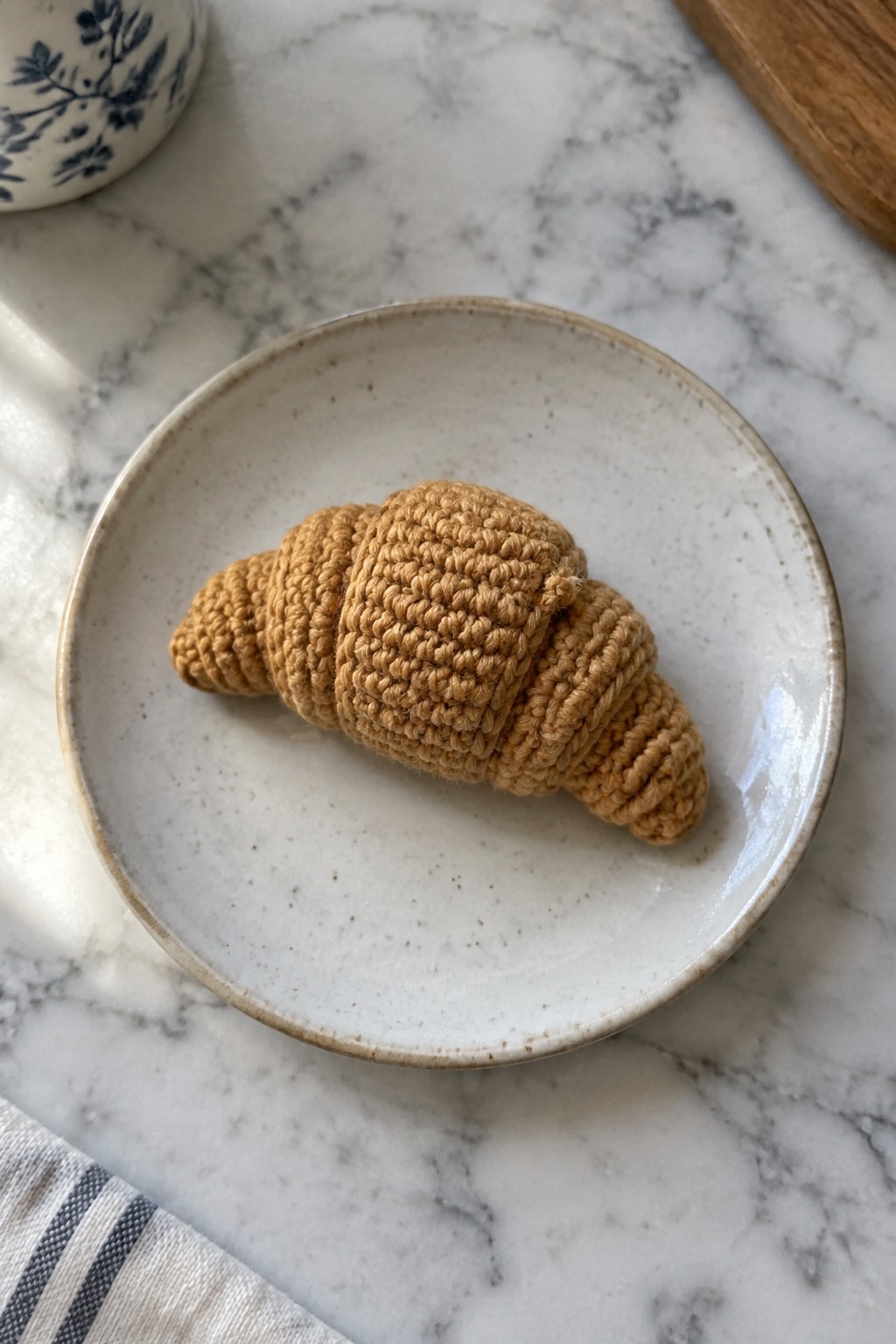 Crocheted tan croissant on speckled white plate atop marble countertop