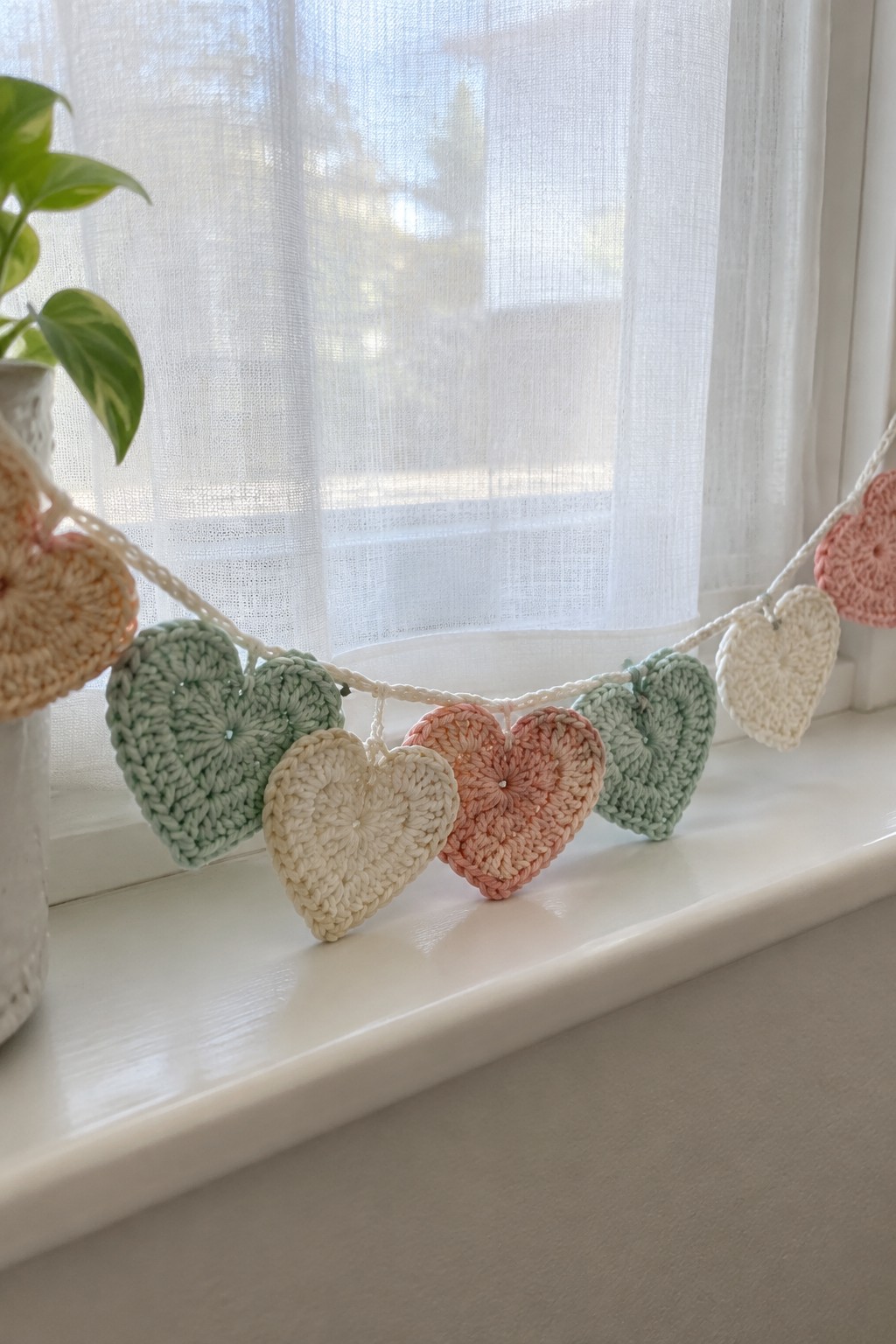 Pastel crocheted heart garland draped across a sunny white windowsill.
