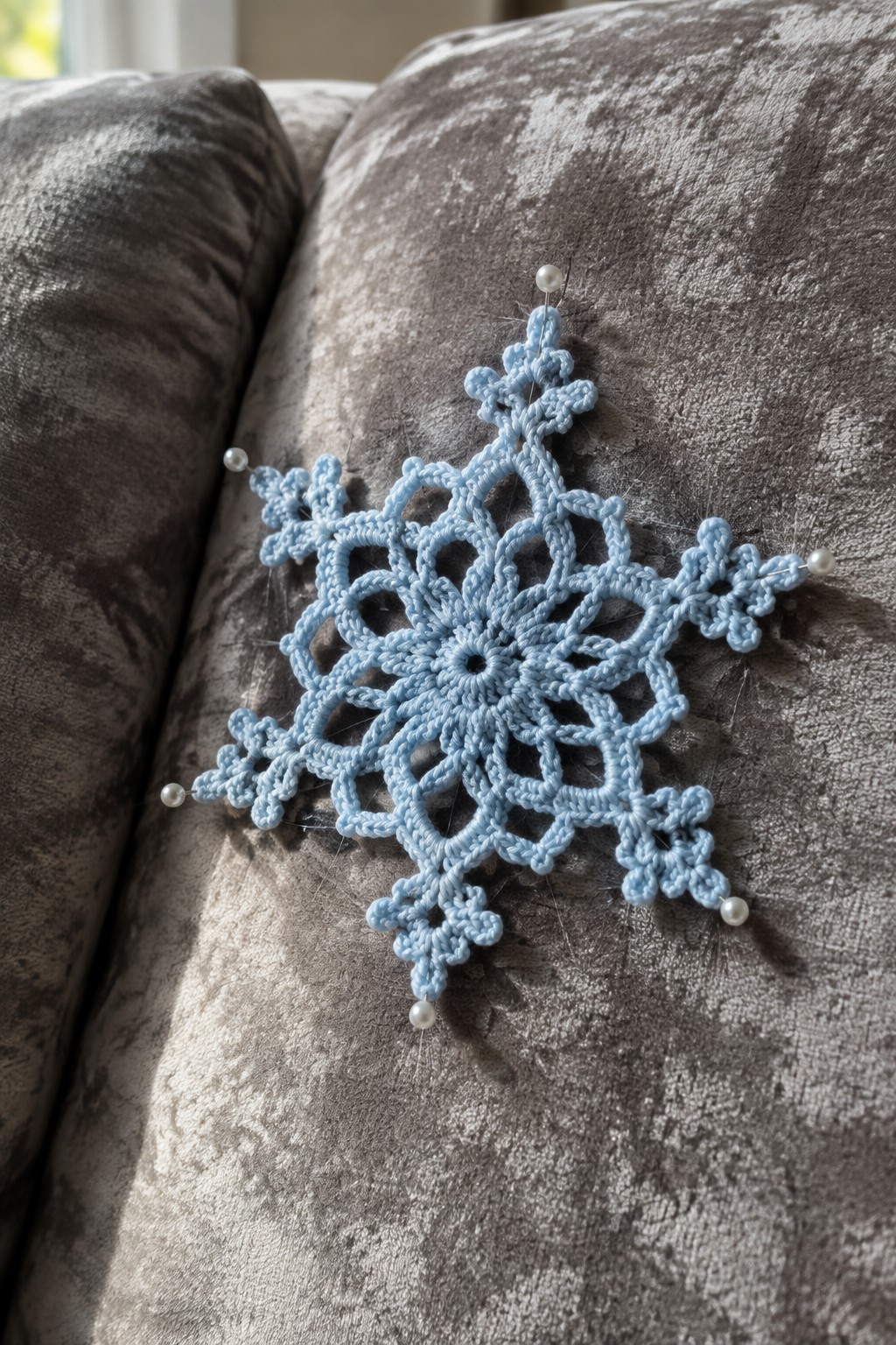 Light blue crocheted snowflake pinned with pearl pins on gray velvet couch