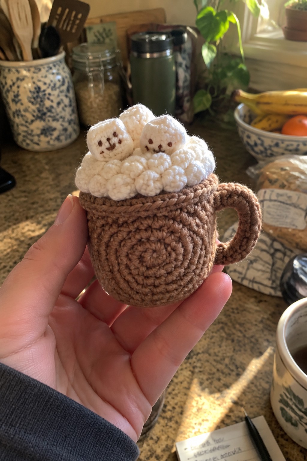 A hand holds a small crocheted brown mug with a handle and white marshmallow shapes on top, some with simple faces.