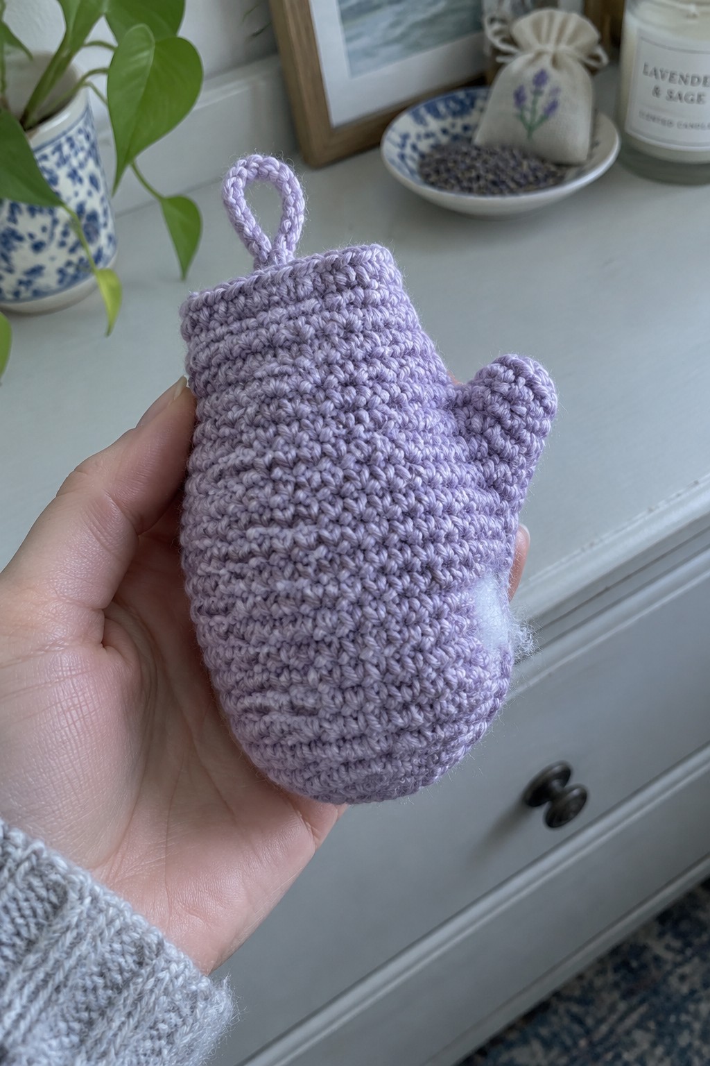 A hand holds a small purple crocheted mitten ornament with a loop at the top.