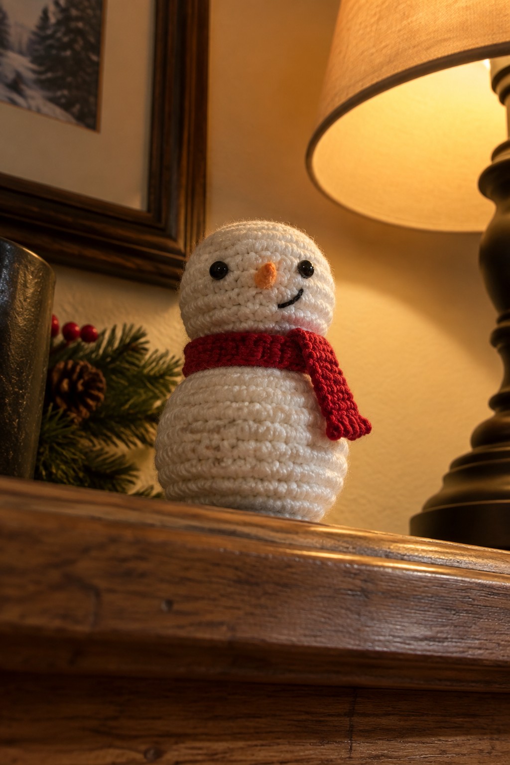 Crocheted white snowman with red scarf on wooden shelf beside glowing lamp