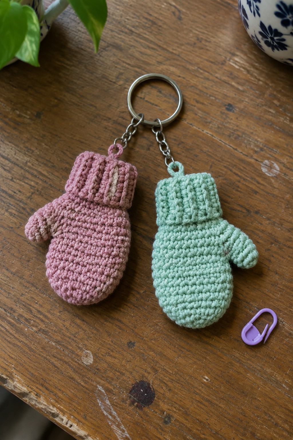 Pink and mint crocheted mitten keychains on wooden table with purple safety pin