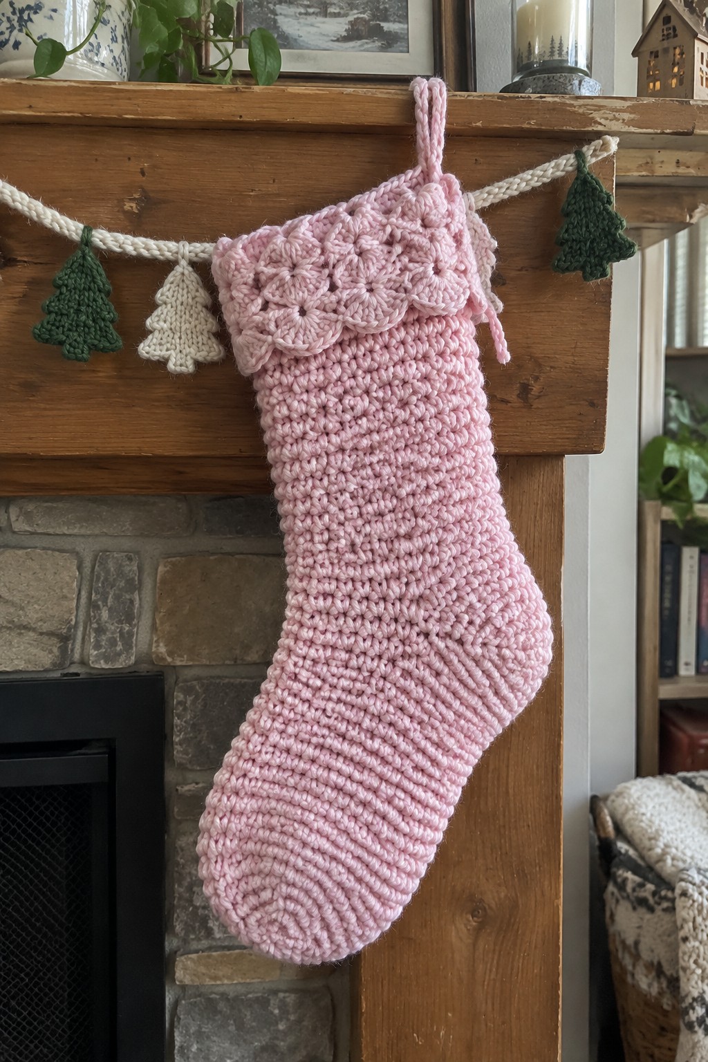 Pink knitted Christmas stocking with floral cuff hangs on wooden mantel beside knitted tree garland