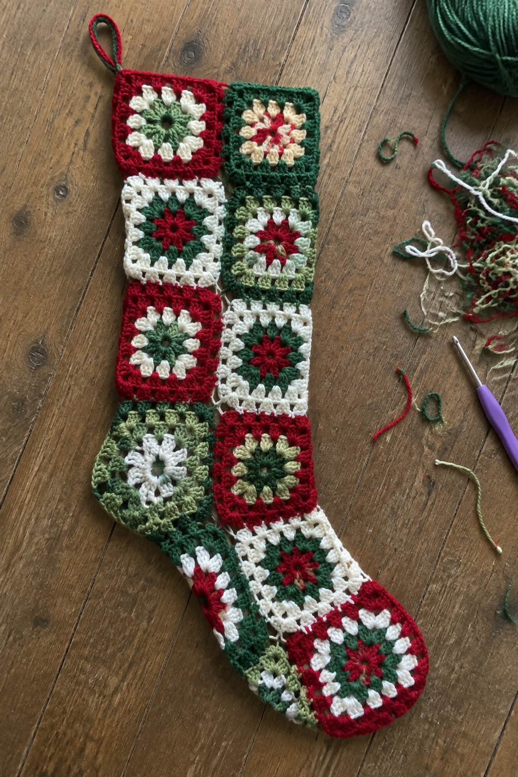 Red, white, and green granny square crochet Christmas stocking with yarn and hook on wood