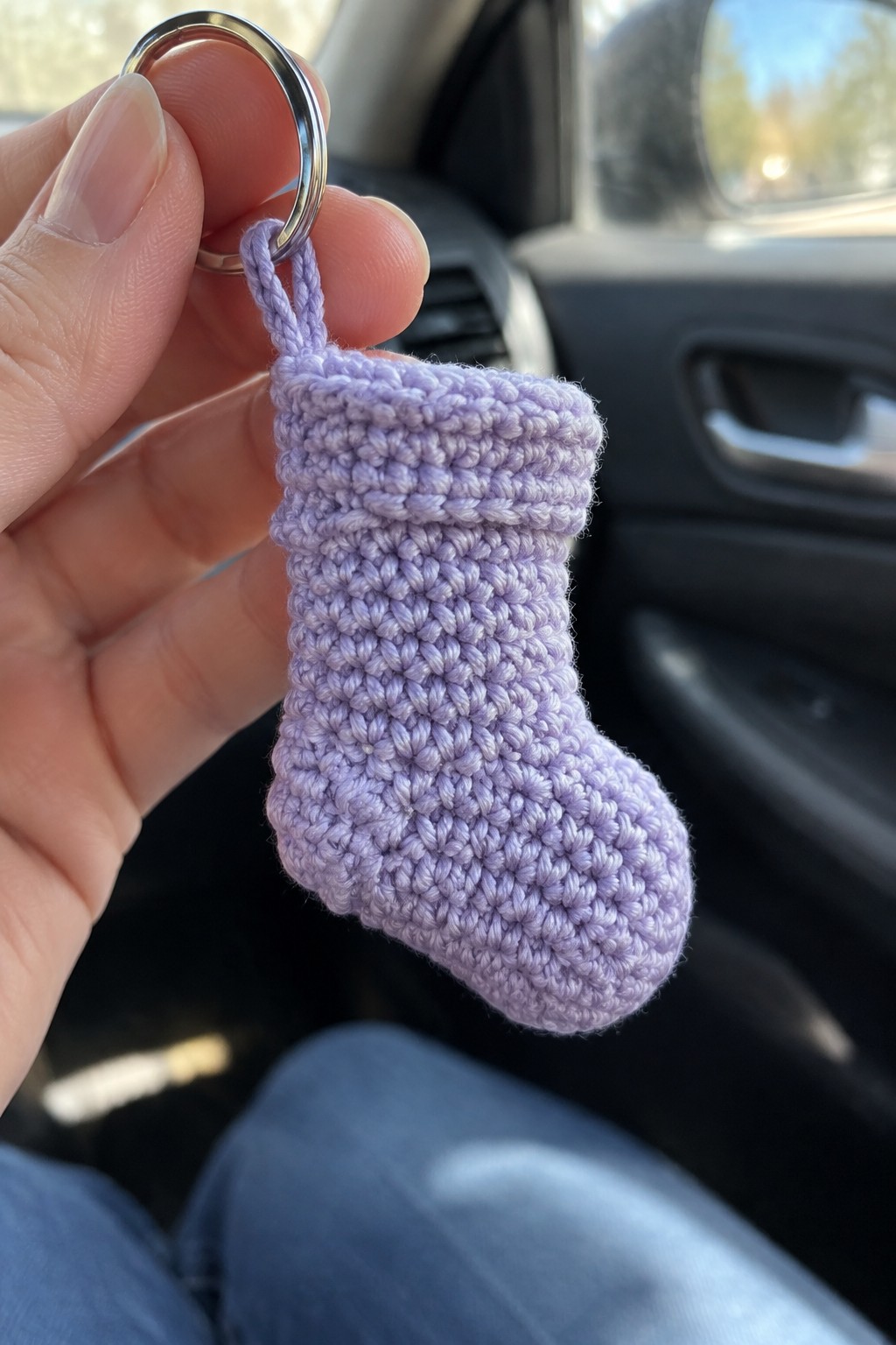 Hand holding tiny purple crocheted stocking keychain with metal ring in car.