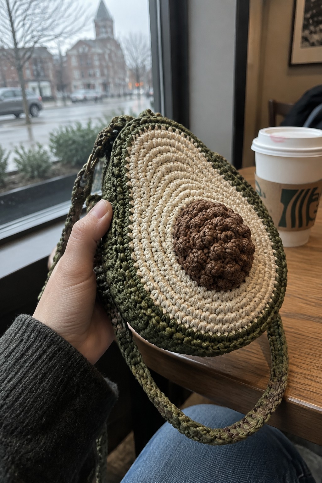 Hand holding green crocheted avocado-shaped bag beside Starbucks cup on wooden table