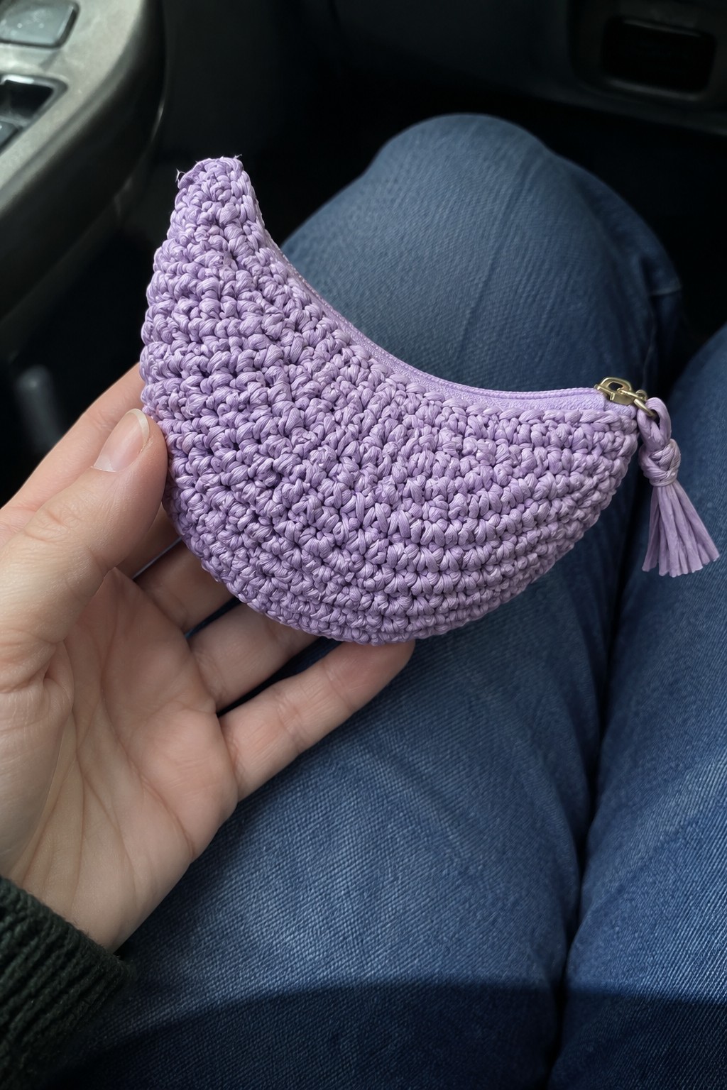 Hand holding lavender crocheted crescent pouch with zipper and tassel on blue jeans