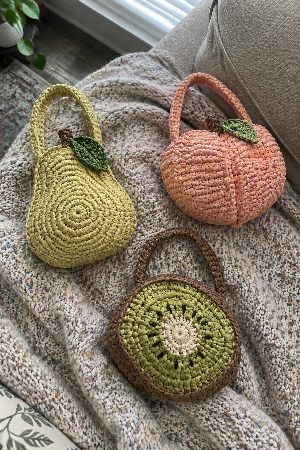 Three crocheted fruit purses—yellow pear, pink peach, green kiwi—on a blanket.