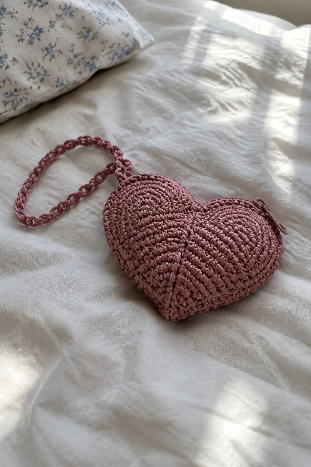 Pink crocheted heart-shaped zippered pouch with braided strap on white bedding