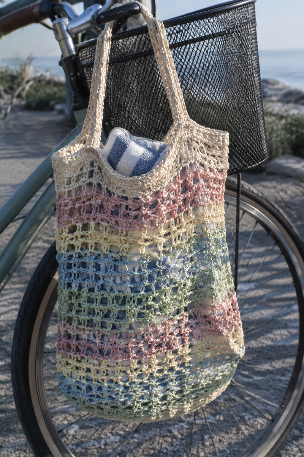 Colorful crocheted mesh tote bag hanging from bicycle handlebars by the sea