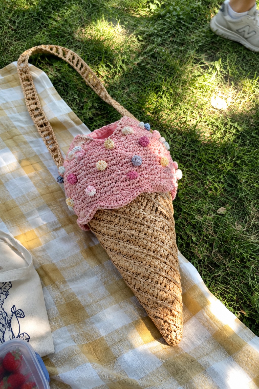 Crocheted raffia ice cream cone bag with tan cone and pink top dotted with colorful pom-poms on a picnic blanket.