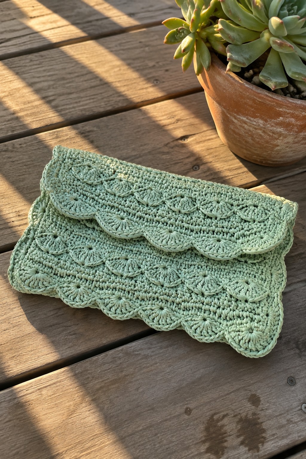 Light green crocheted clutch with scalloped edges on wooden table beside succulent plant
