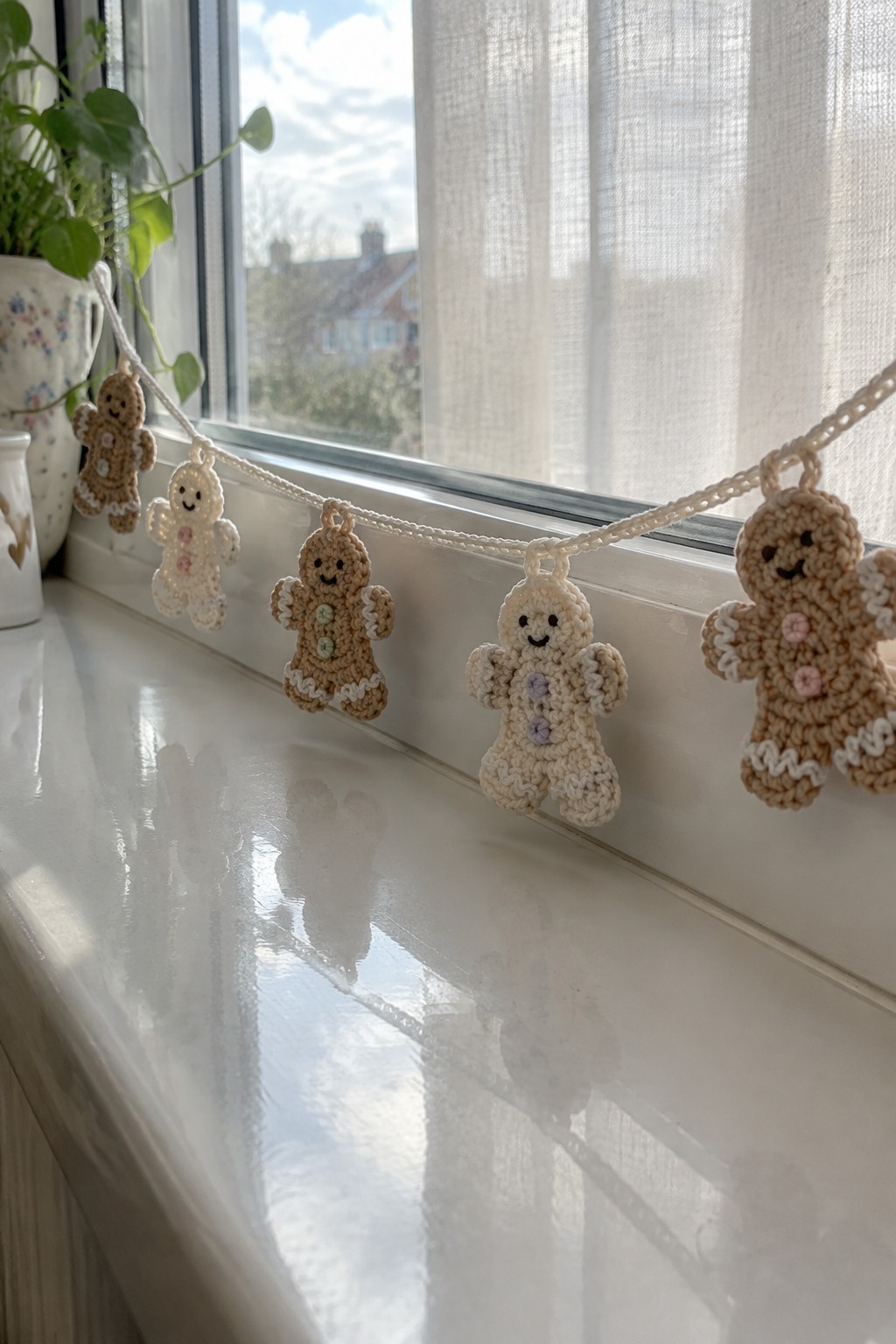 Crocheted gingerbread men in brown and cream yarn are strung together on a white string and draped across a windowsill.