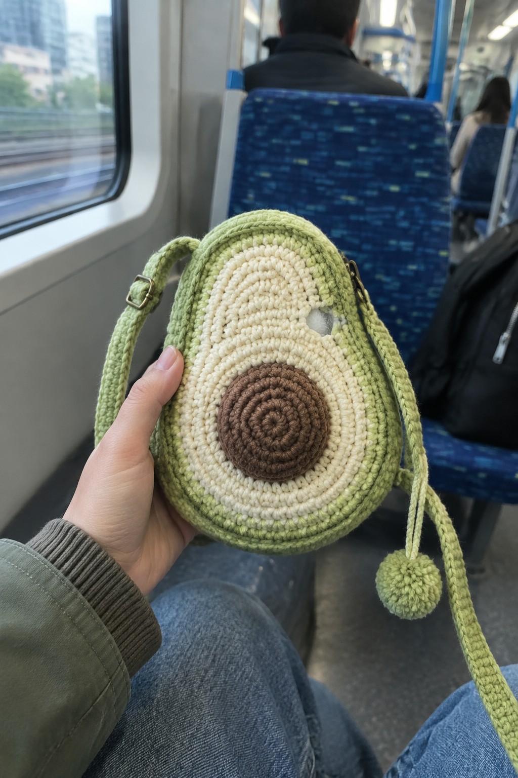 Hand holds crocheted avocado bag on lap inside blue train seat
