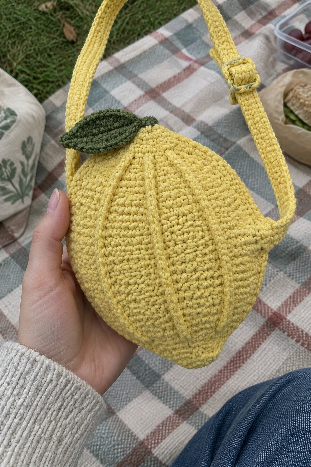 Hand holding yellow crocheted lemon bag with green leaf on checkered picnic blanket