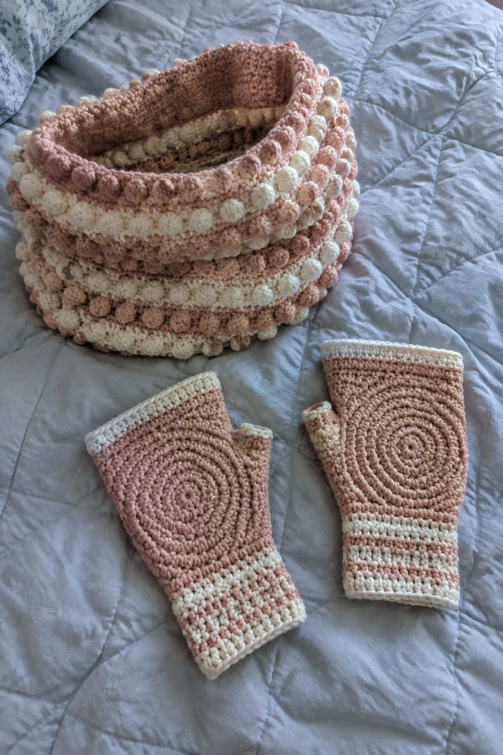Pink and white crocheted cowl with bobble texture beside spiral mittens on quilt.