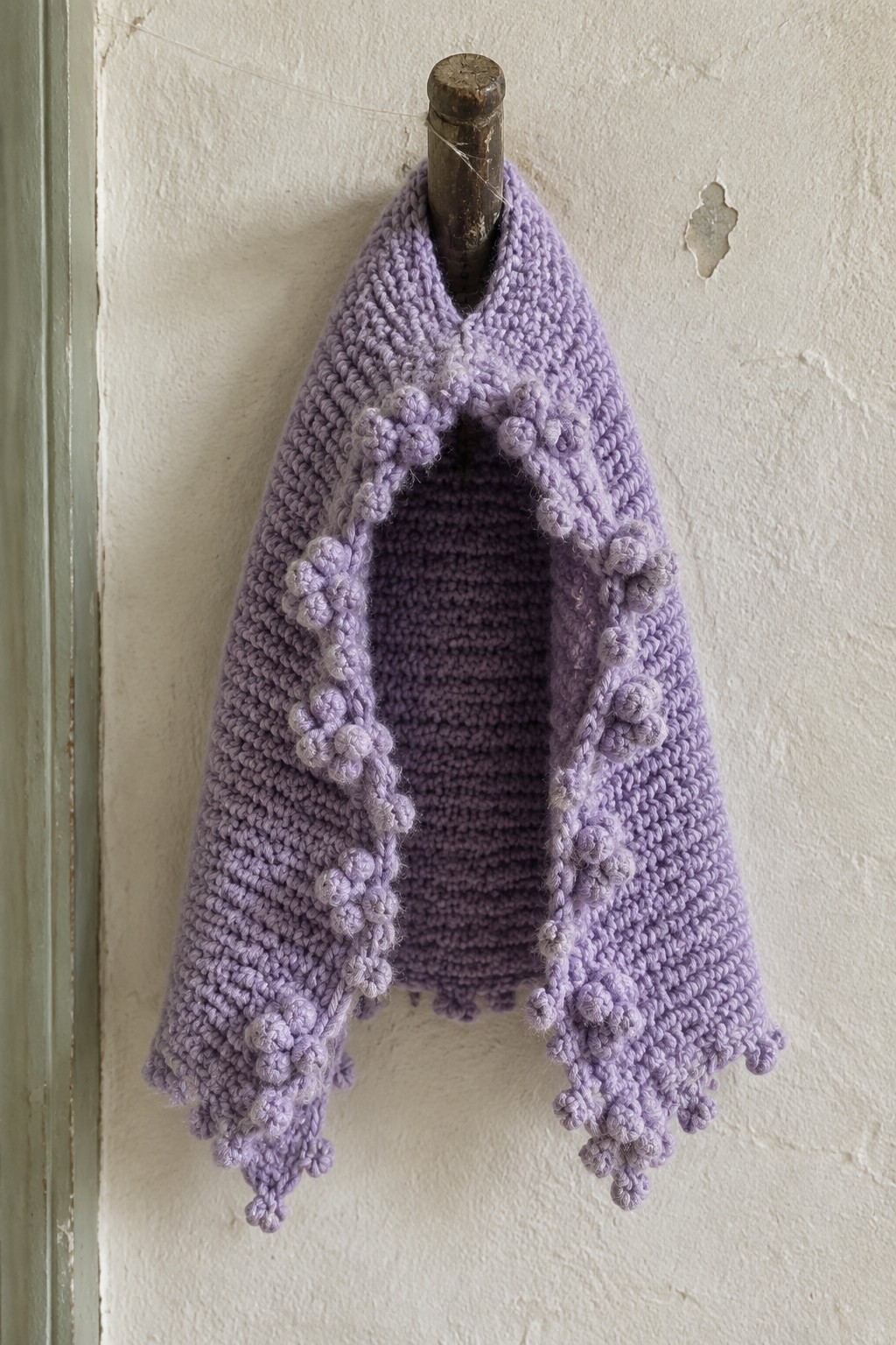 Lavender knitted shawl with pom-pom trim hanging on wooden peg against white wall