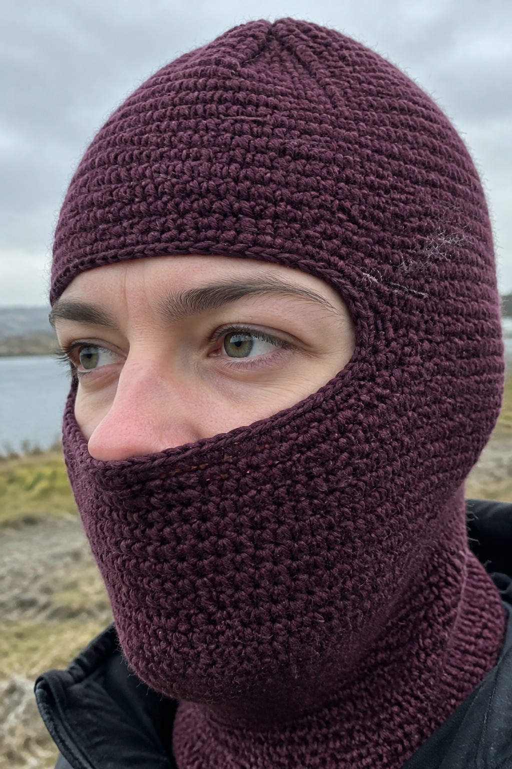 Person in maroon knitted balaclava with green eyes outdoors near water