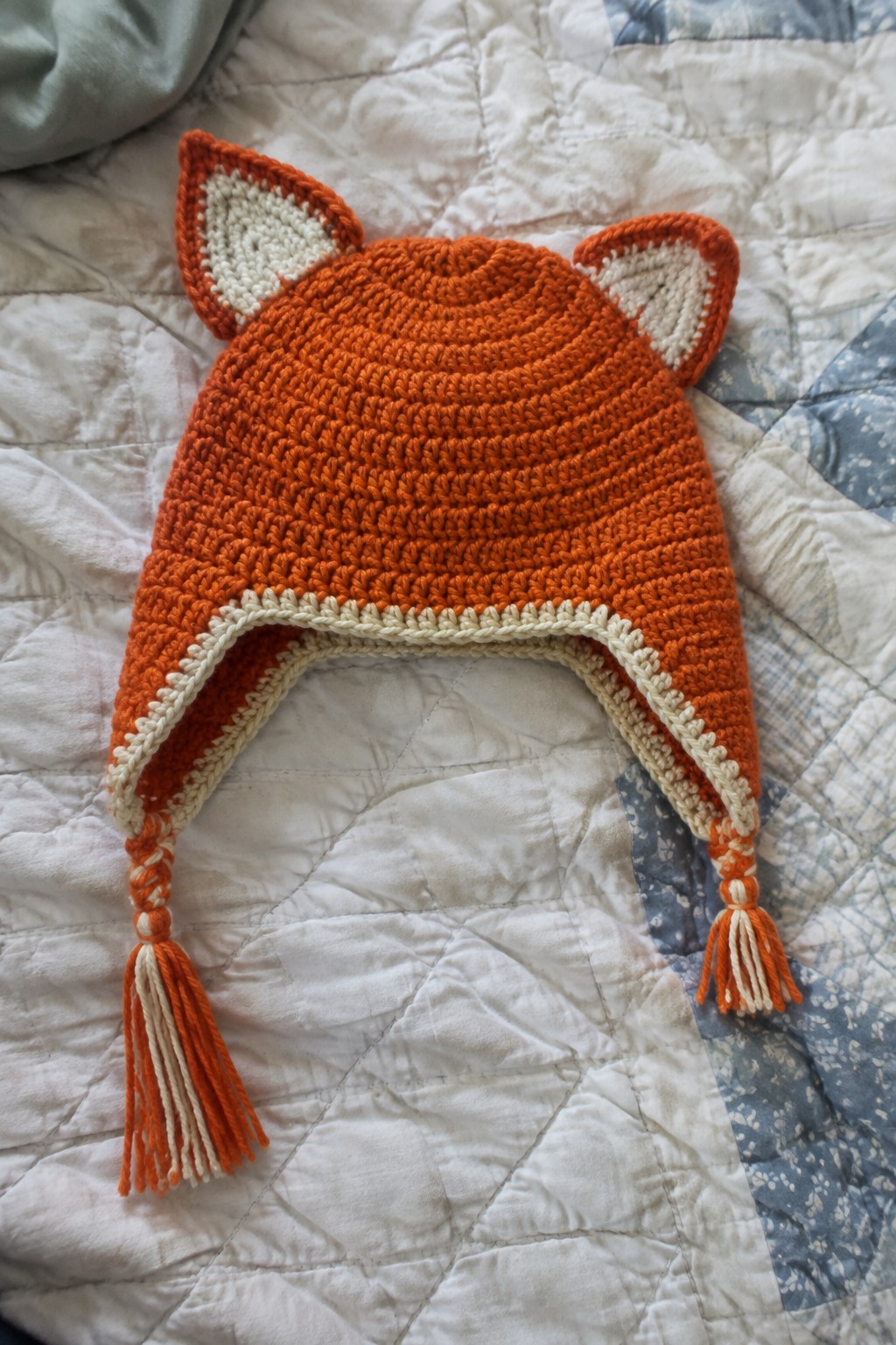 Crocheted orange fox hat with white ears and tassel ties on quilt.
