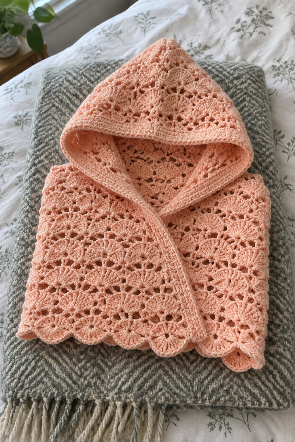 Peach crocheted hooded poncho on gray herringbone fringed blanket atop bed