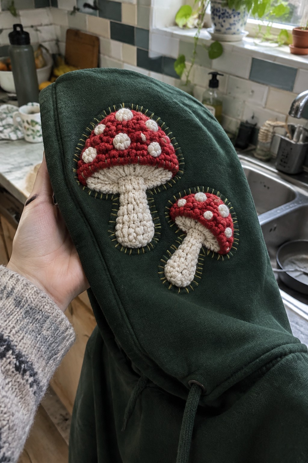 Green hoodie with two crocheted red-and-white mushrooms held in a kitchen