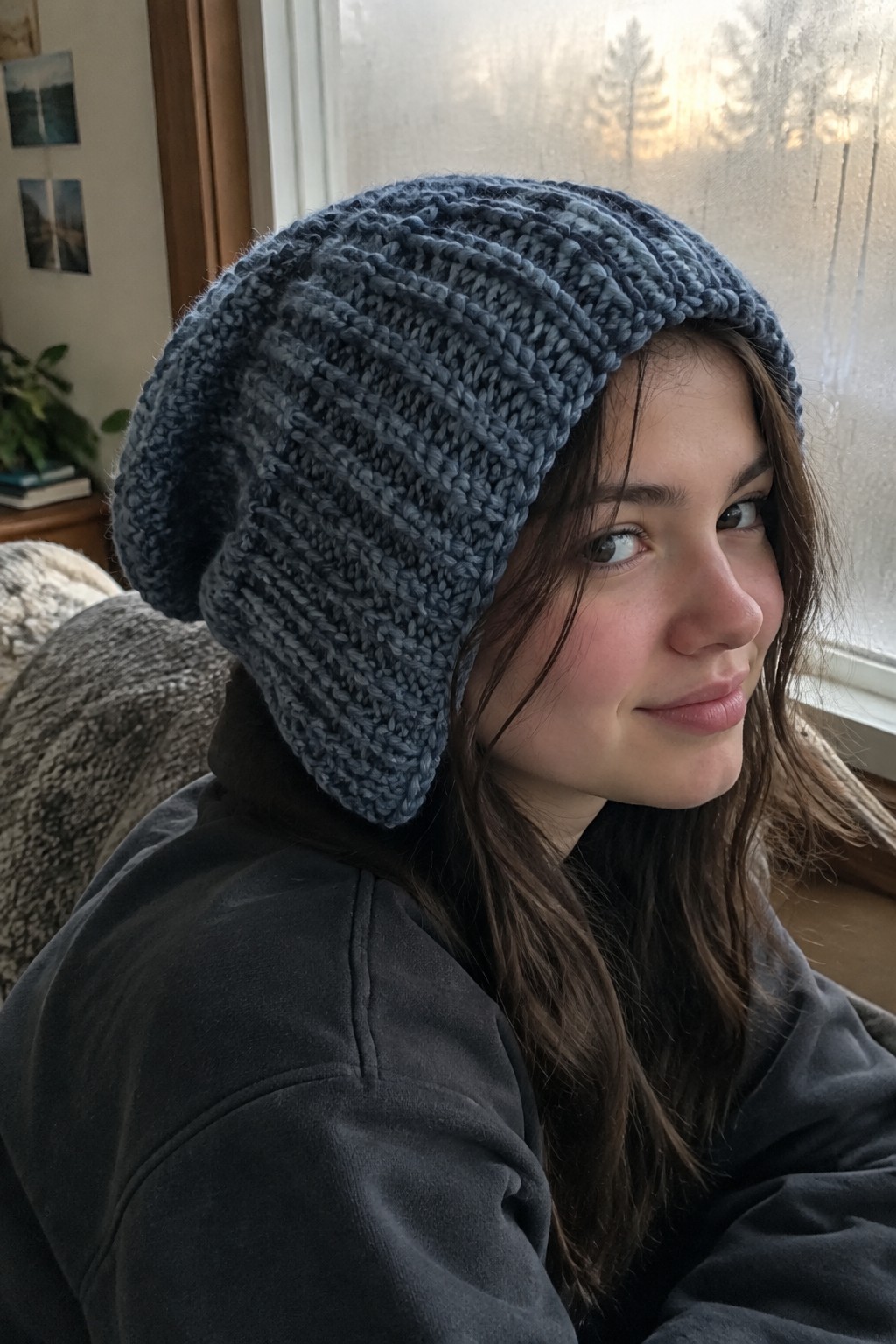 Young woman in chunky blue knit hat and dark hoodie by frosted window