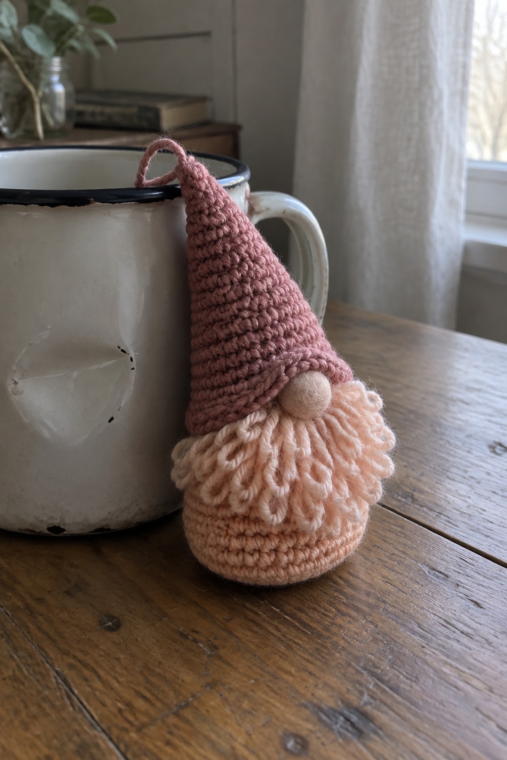 Pink crocheted gnome with peach beard leaning on white enamel mug on wood table