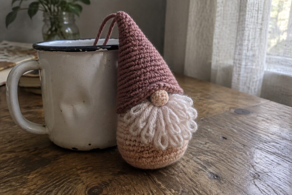 23 Quick Christmas Crochet Projects for Last-Minute Holiday Making Pink crocheted gnome with peach beard leaning on white enamel mug on wood table