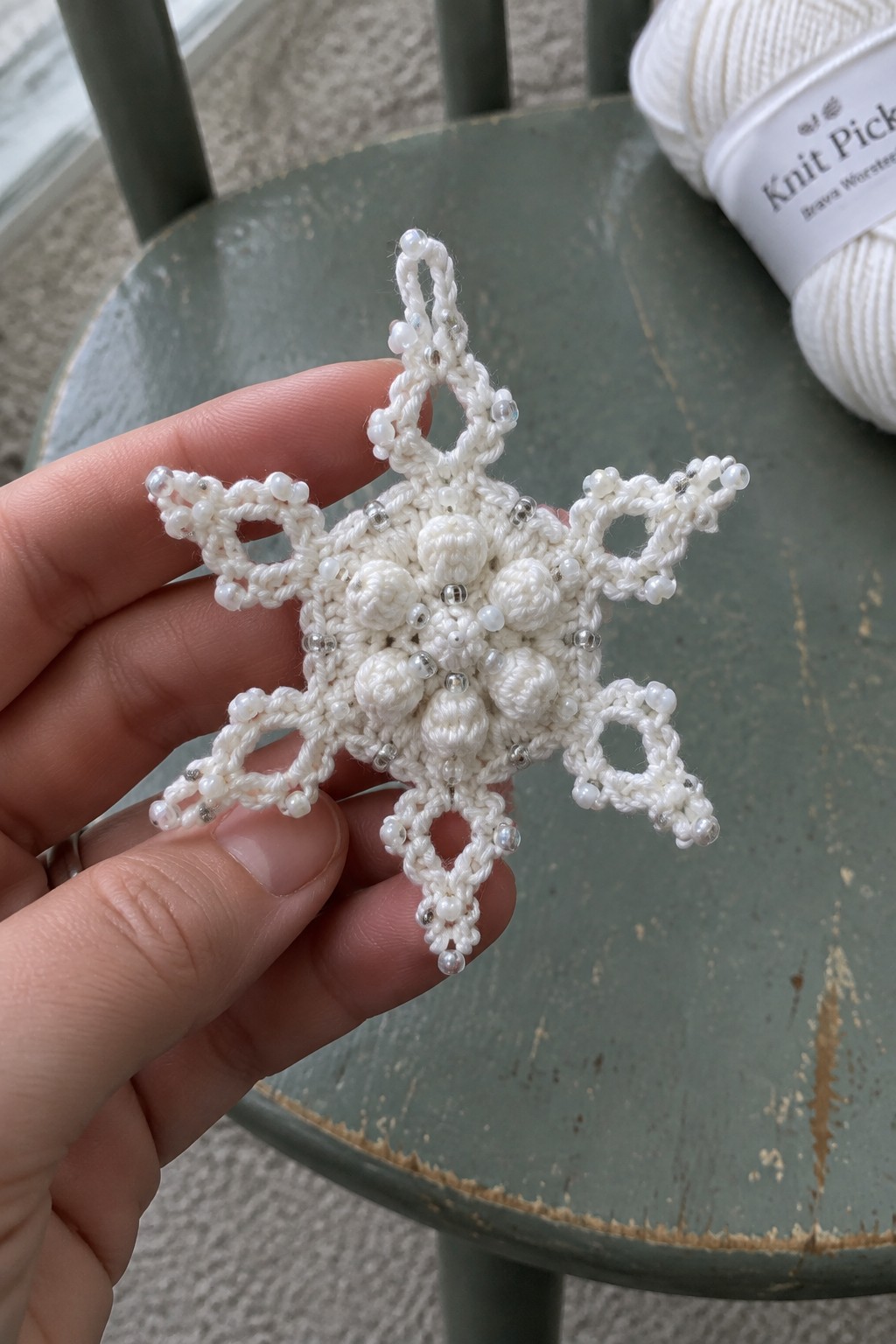 Hand holding white beaded crocheted snowflake on green table with yarn skein