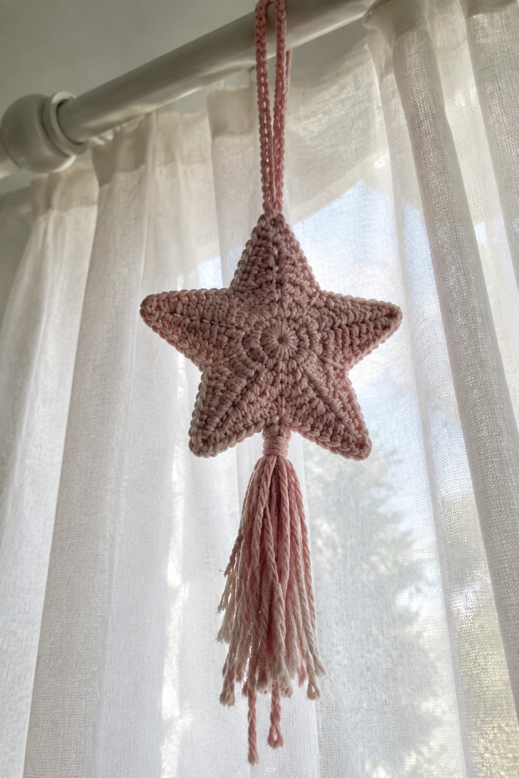 Pink crocheted star ornament with tassel hanging from white curtain rod.