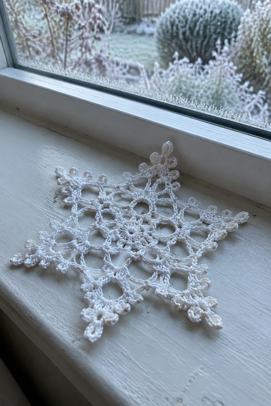 White crocheted snowflake doily on frosty windowsill with snowy garden view outside.