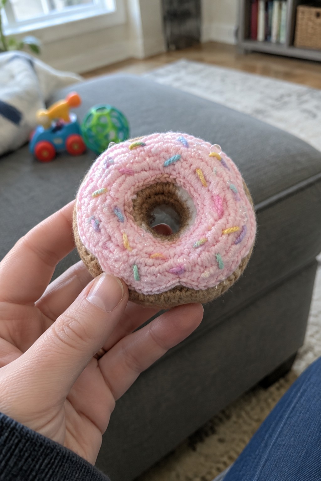 Hand holding pink crocheted donut toy with colorful sprinkles on gray ottoman