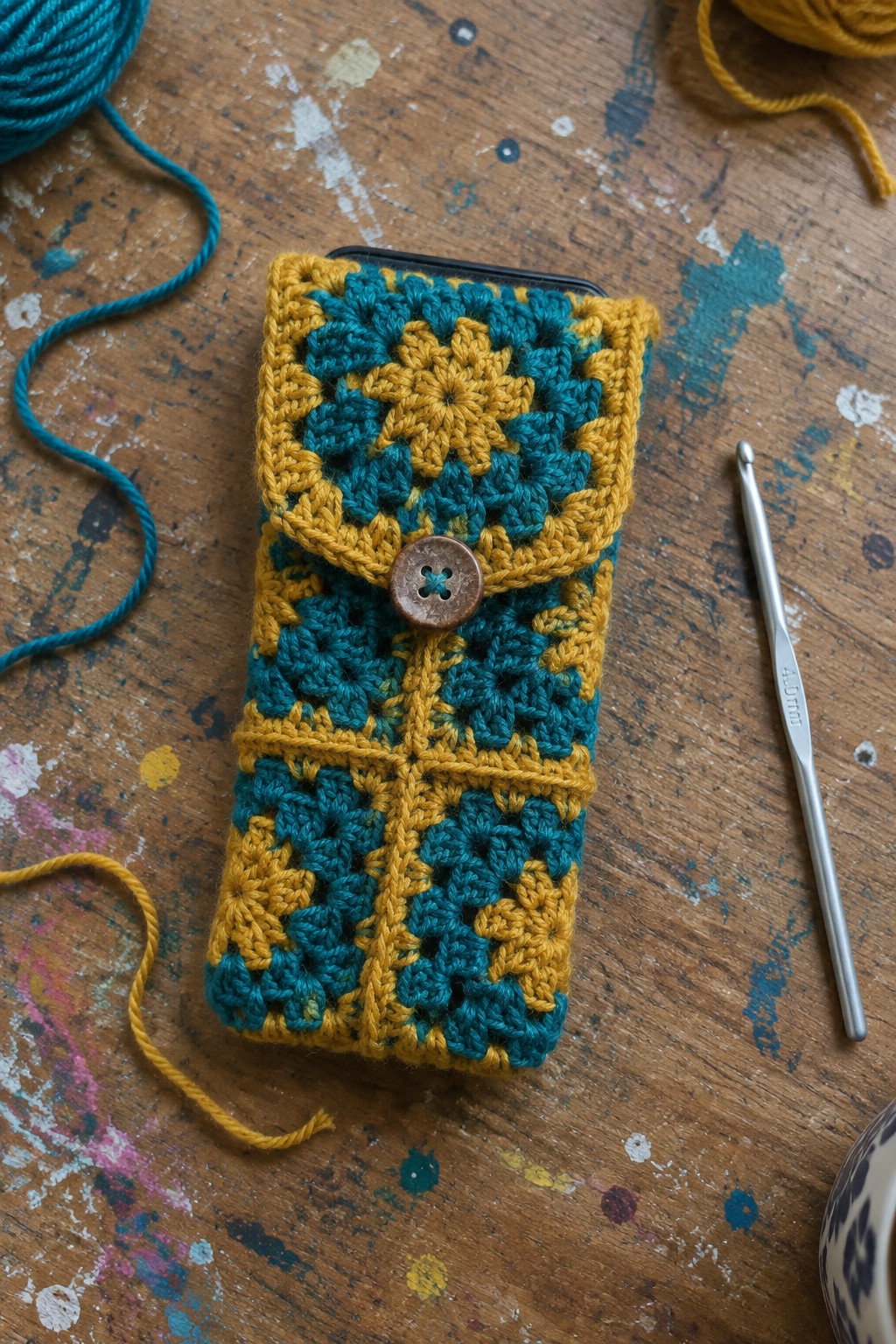 Teal and yellow granny square crocheted phone pouch with button on wooden table