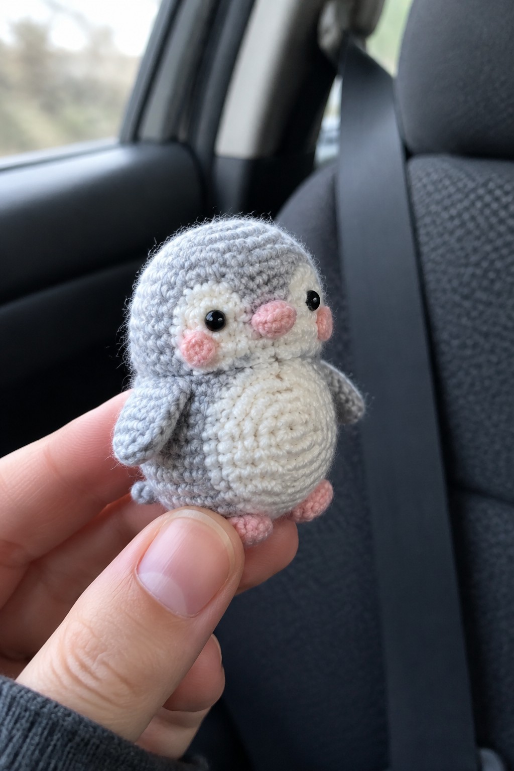 Hand holding small gray crocheted penguin with pink cheeks inside car