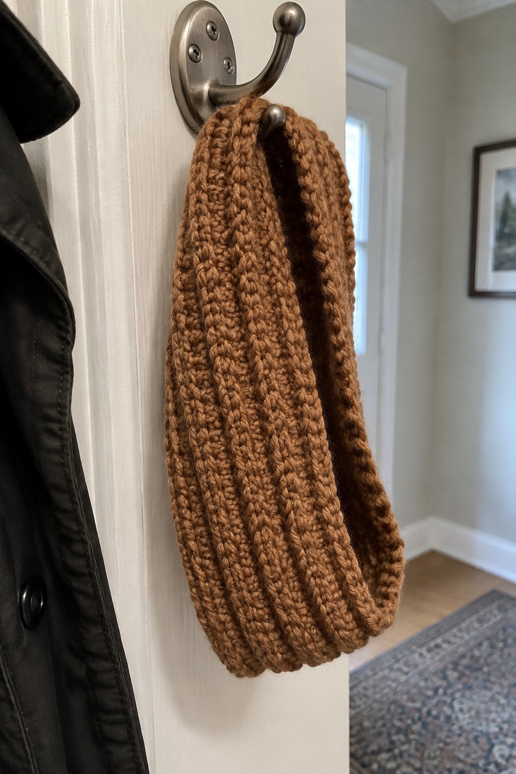 Brown knitted scarf hanging on metal hook beside black jacket on white door
