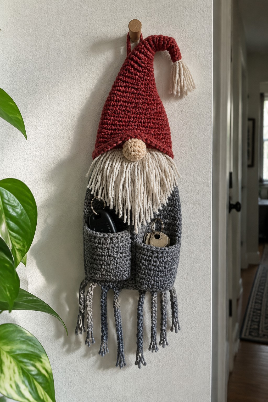 Crocheted gnome wall hanging with red hat, white beard, and gray pockets holding keys on a wooden hook.