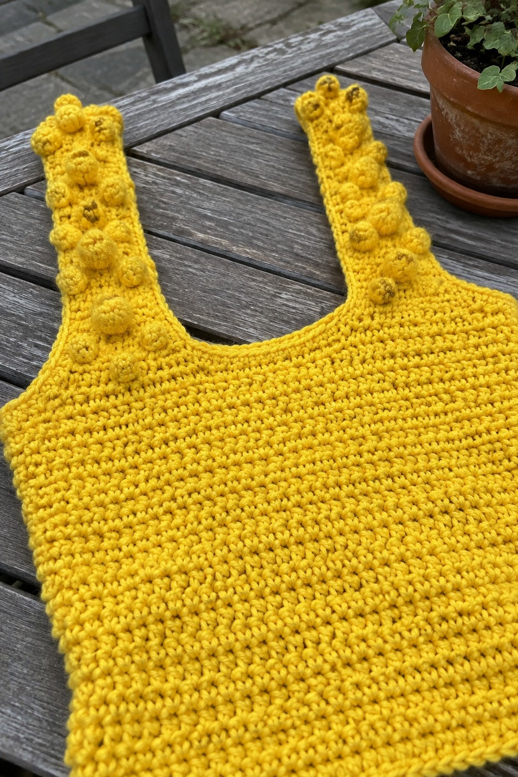 Yellow crocheted tank top with bobble straps on wooden table beside potted plant