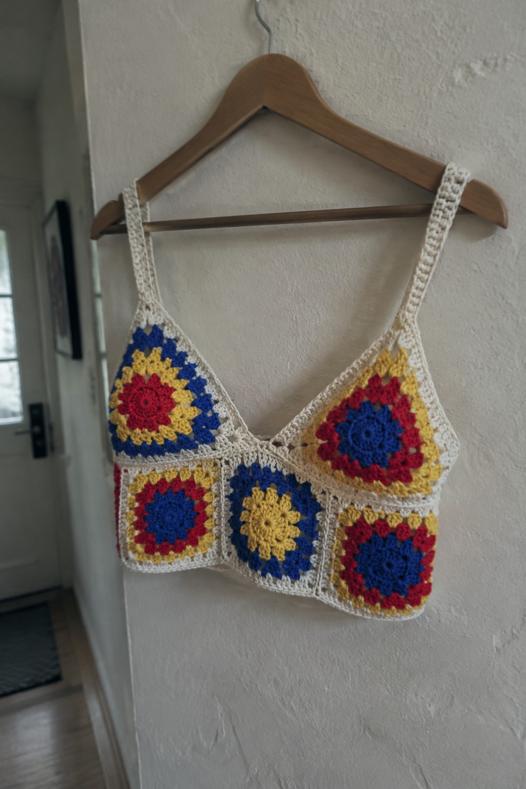 Colorful crocheted granny square crop top hanging on wooden hanger against white wall.