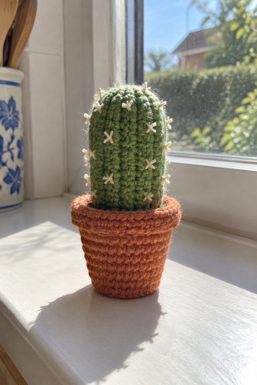 A green crocheted cactus with white star accents sits inside an orange crocheted pot on a white windowsill.