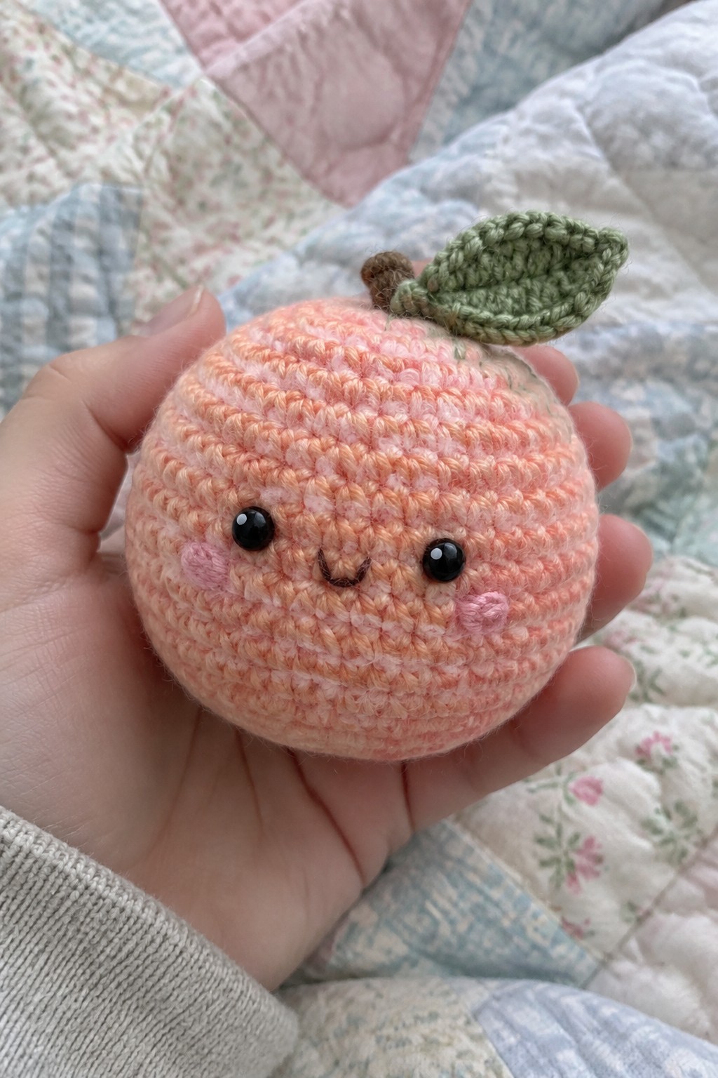 Hand holding crocheted peach amigurumi with smiling face, pink cheeks, and green leaf.