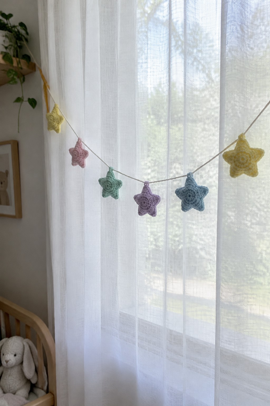 A string of crocheted pastel stars in yellow, pink, mint, lavender, and blue hangs across a window with sheer white curtains.