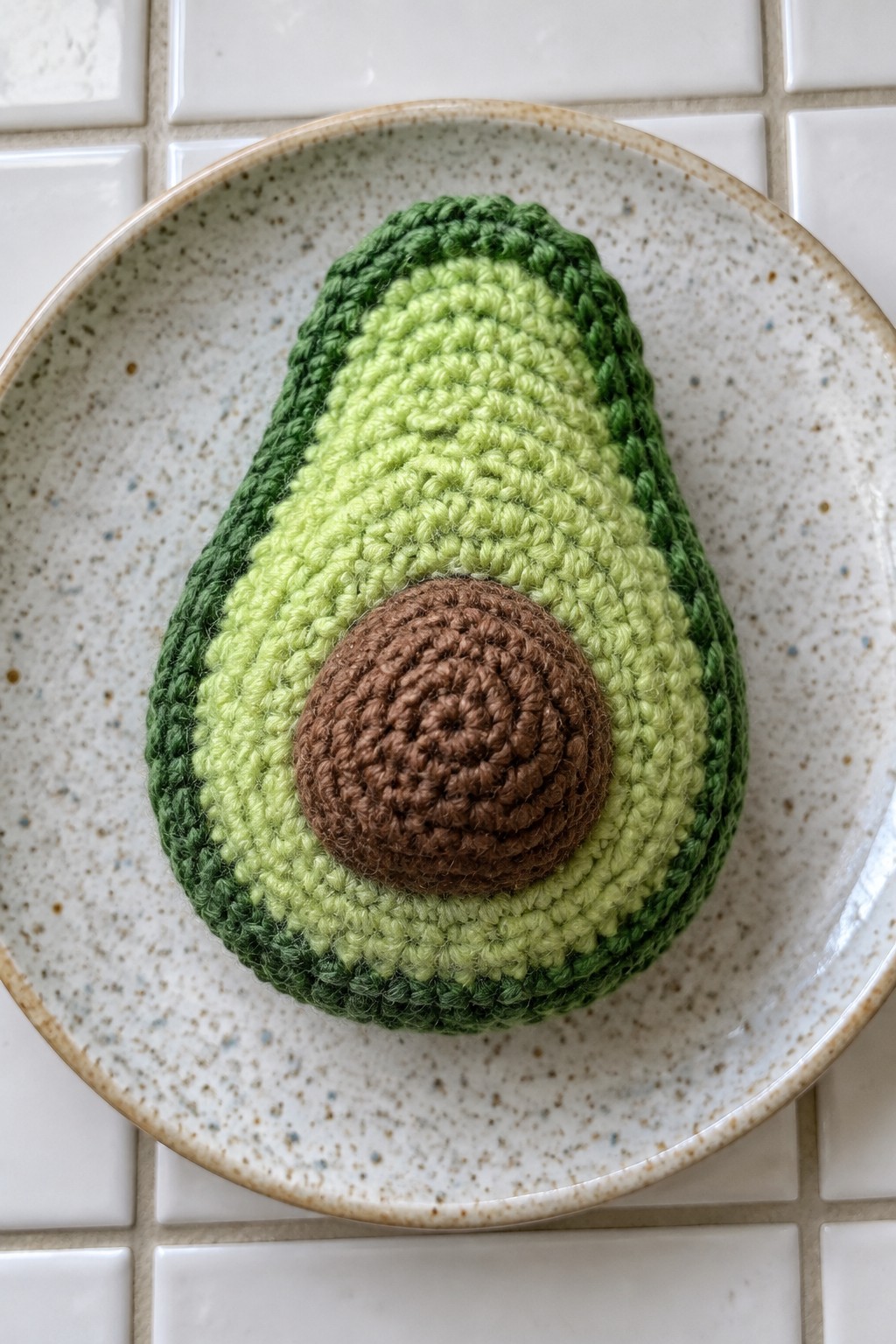 Crocheted avocado with dark green rind, light flesh, and brown pit on speckled plate