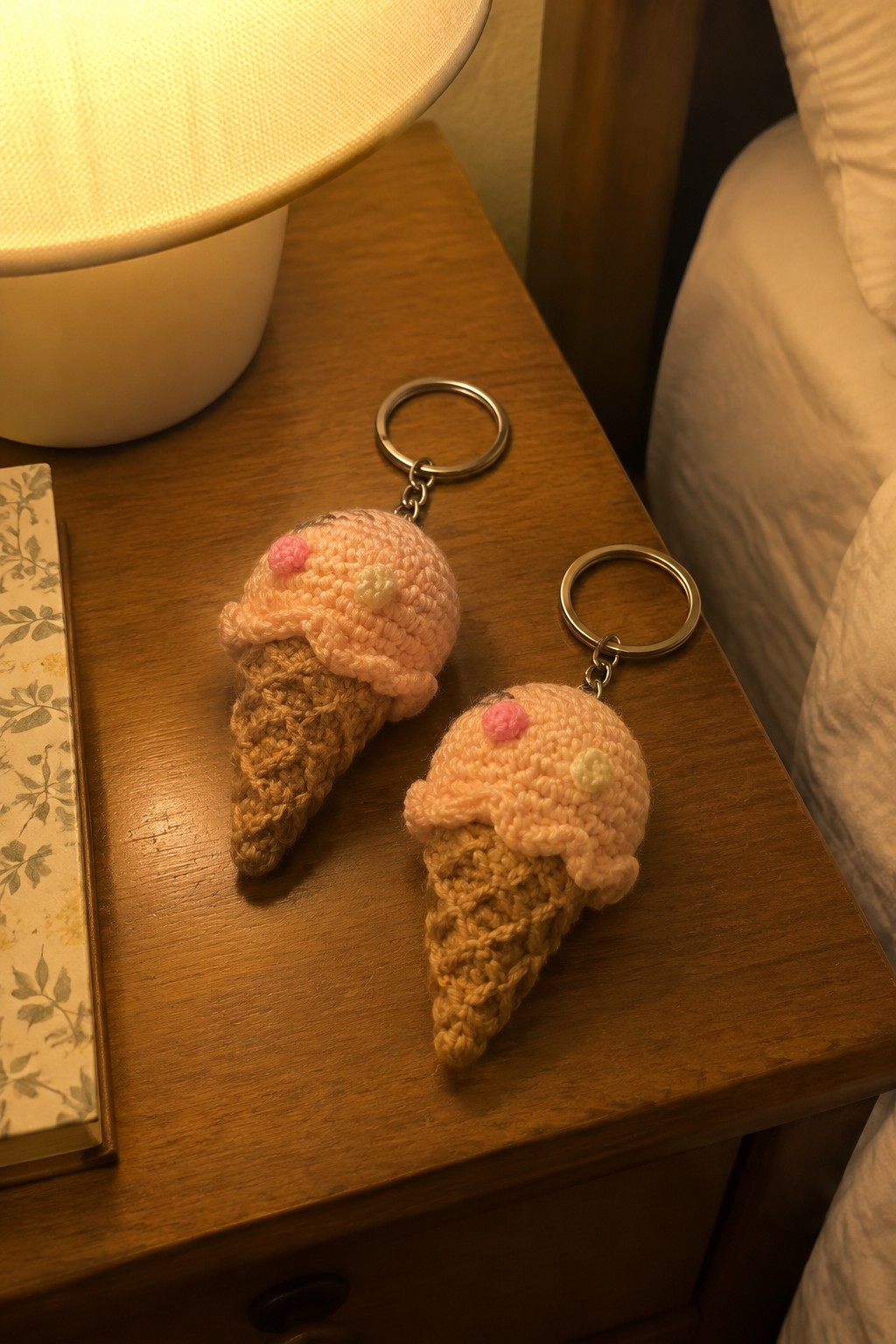 Two crocheted ice cream cone keychains on a wooden nightstand beside a lamp and book