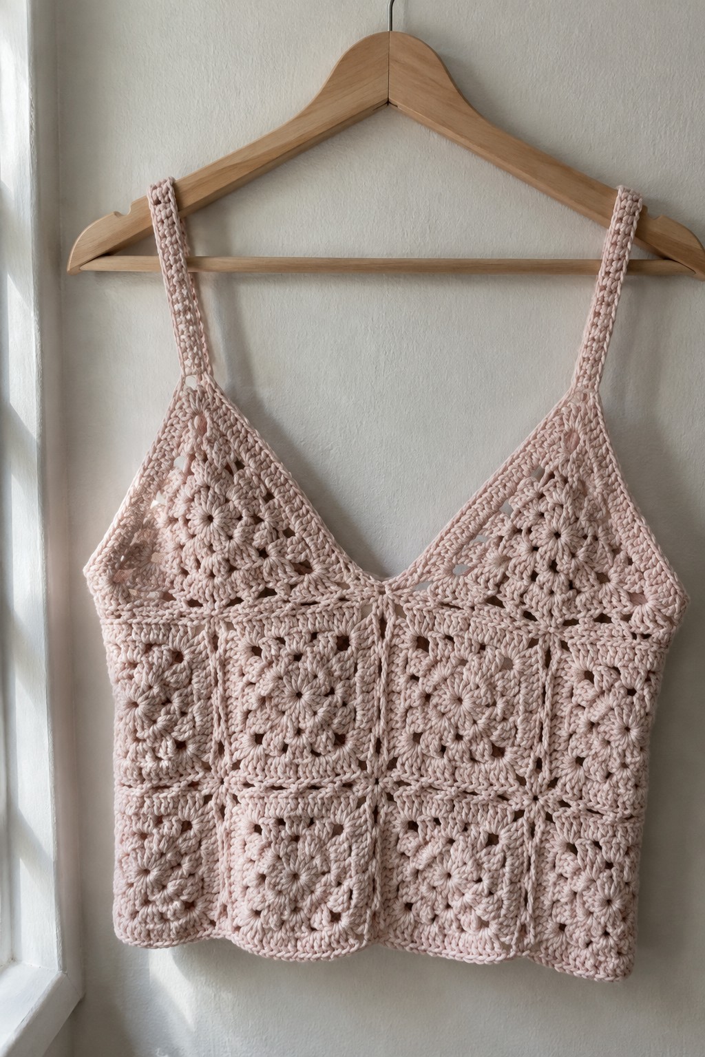 Soft pink granny square crochet camisole on wooden hanger against wall.